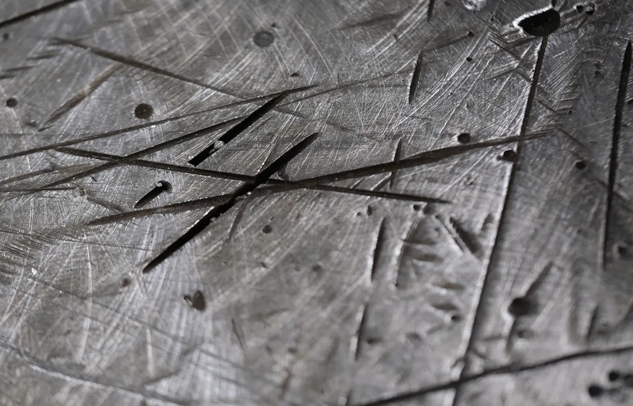 Macro shot of a worn steel plate covered in crossing gouges, scratches, and small pits in cool charcoal tones